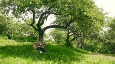 Parktaki bir ağacın altında gitarını tıngırdatan bir müzisyenin sakin sahnesi. Nazik melodiler, bir adamın bir ağacın altında gitar çalması gibi havayı doldurur. Rahatlatıcı an: adam ağacın gölgesinde gitar çalıyor. Adam ağacın altında akustik gitar çalıyor..
