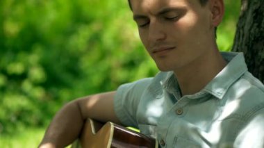 Parktaki bir ağacın altında gitarını tıngırdatan bir müzisyenin sakin sahnesi. Nazik melodiler, bir adamın bir ağacın altında gitar çalması gibi havayı doldurur. Rahatlatıcı an: adam ağacın gölgesinde gitar çalıyor. Adam ağacın altında akustik gitar çalıyor..