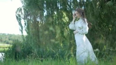 Nehir kıyısındaki çimlerin üzerinde zarif bir şekilde duran beyaz, eski moda elbiseli kadın. Yeşil çimenler ve ağaçların arasına işlenmiş ulusal nakışlı elbiseler içinde zarif bir bayan. Çimenli bir tarlada gülümseyen, beyaz elbiseli, huzurlu Ukraynalı kadın. Beyaz elbiseli kadın etrafını sardı 