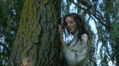 Nehir kıyısındaki çimlerin üzerinde zarif bir şekilde duran beyaz, eski moda elbiseli kadın. Yeşil çimenler ve ağaçların arasına işlenmiş ulusal nakışlı elbiseler içinde zarif bir bayan. Çimenli bir tarlada gülümseyen, beyaz elbiseli, huzurlu Ukraynalı kadın. Beyaz elbiseli kadın etrafını sardı 