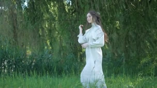 川岸の草の上に優雅に立っている白いヴィンテージのドレスの女性 緑の草と木の間の国の刺繍されたドレスのエレガントな女性 白い服を着たセレン ウクライナ人女性が草原で微笑んでいる 包囲された白衣の女性 — ストック動画