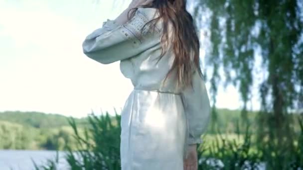 川岸の草の上に優雅に立っている白いヴィンテージのドレスの女性 緑の草と木の間の国の刺繍されたドレスのエレガントな女性 白い服を着たセレン ウクライナ人女性が草原で微笑んでいる 包囲された白衣の女性 — ストック動画