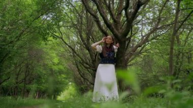 Doğanın güzelliği içinde poz veren geleneksel beyaz elbiseli bir kadın. Ukraynalı beyaz elbiseli kadın sakin bir orman ortamında zarafetle ayakta duruyor. Etrafı yemyeşil yemyeşil bir ormanla çevrili beyaz elbiseli bir kadın. Zarif bir bayan. 