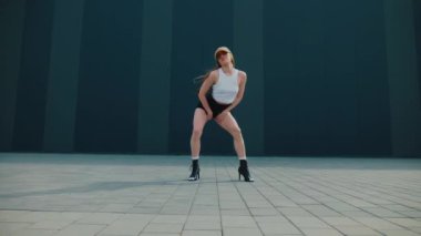 Binanın önünde dans eden bir kadın. Yavaş çekim. Beyaz bluzlu ve siyah şortlu bir kadın binanın önünde poz veriyor. Mimari arka planda sokak dansı pozu. Modern bir binanın yanında şehir dansı havası var. Şık bir kadın.