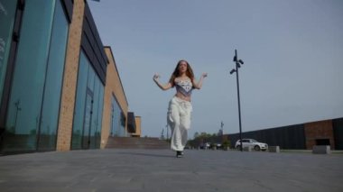 Kadın şehir binasının önünde zarif bir şekilde yüksek topuklularla dans ediyor. Enerjik kadın dışarıda dans gösterisi yapıyor. Dansçı şehrin arka planında neşe ifade ediyor. Kadın dansçı şehir planıyla büyülenir. Neşeli kadın dans yeteneklerini sergiliyor. 