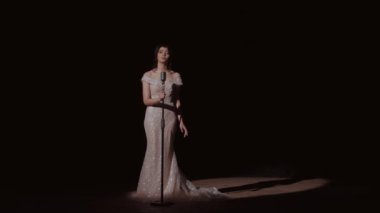 Gece kıyafetli kadın mikrofonun yanında tek başına duruyor. Kız mikrofona konuşuyor. Genç bir kız büyük, karanlık bir salonda şarkı söylüyor. Yumuşak ışıkta beyaz elbiseler içinde zarif bir gelin. Parlak arkaplanı olan gelinlikli bir kadın. Gelin güzeli.