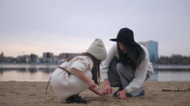 Anne ve kızı su kenarında birbirlerine sarılıp duruyorlar. Kadın ve çocuğun manzaranın tadını çıkardığı plaj sahnesi. Kadın ve kız gülümsüyor ve sahilde duruyor, denize bakıyor. Kadın ve küçük kız sahilde ufka bakıyor. Kumlu sahilde aile zamanı