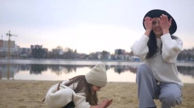 Anne ve kızı su kenarında birbirlerine sarılıp duruyorlar. Kadın ve çocuğun manzaranın tadını çıkardığı plaj sahnesi. Kadın ve kız gülümsüyor ve sahilde duruyor, denize bakıyor. Kadın ve küçük kız sahilde ufka bakıyor. Kumlu sahilde aile zamanı