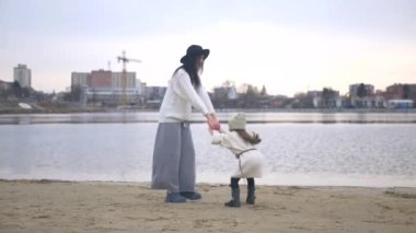 Anne ve kızı su kenarında birbirlerine sarılıp duruyorlar. Kadın ve çocuğun manzaranın tadını çıkardığı plaj sahnesi. Kadın ve kız gülümsüyor ve sahilde duruyor, denize bakıyor. Kadın ve küçük kız sahilde ufka bakıyor. Kumlu sahilde aile zamanı