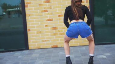 Kot şortlu ve siyah bluzlu bir kadın şehir binasının önünde yüksek topuklularla zarif bir şekilde dans ediyor. Yavaş çekim. Enerjik kadın dışarıda dans gösterisi yapıyor. Dansçı şehrin arka planında neşe ifade ediyor. Kadın dansçı şehir manzarasına hayran.