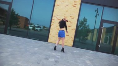 Kot şortlu ve siyah bluzlu bir kadın şehir binasının önünde yüksek topuklularla zarif bir şekilde dans ediyor. Yavaş çekim. Enerjik kadın dışarıda dans gösterisi yapıyor. Dansçı şehrin arka planında neşe ifade ediyor. Kadın dansçı şehir manzarasına hayran.