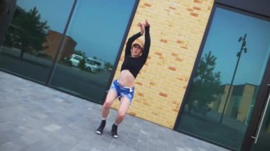 Kot şortlu ve siyah bluzlu bir kadın şehir binasının önünde yüksek topuklularla zarif bir şekilde dans ediyor. Yavaş çekim. Enerjik kadın dışarıda dans gösterisi yapıyor. Dansçı şehrin arka planında neşe ifade ediyor. Kadın dansçı şehir manzarasına hayran.