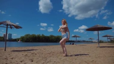 Bachata, salsa ve kizomba yapan bir kadın. Sahilde, mavi gökyüzünün arka planında. Gülümseyen kadın çıplak ayakla poz verir ve sıcak kumda baştan çıkarıcı bir şekilde dans eder. Plaj sahnesinde kızın keyfi yerinde. Şortlu ve tişörtlü güzel bayan poz veriyor.