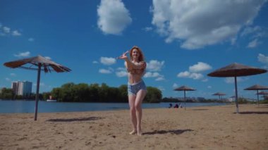 Bachata, salsa ve kizomba yapan bir kadın. Sahilde, mavi gökyüzünün arka planında. Gülümseyen kadın çıplak ayakla poz verir ve sıcak kumda baştan çıkarıcı bir şekilde dans eder. Plaj sahnesinde kızın keyfi yerinde. Şortlu ve tişörtlü güzel bayan poz veriyor.