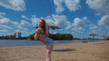 Bachata, salsa ve kizomba yapan bir kadın. Sahilde, mavi gökyüzünün arka planında. Gülümseyen kadın çıplak ayakla poz verir ve sıcak kumda baştan çıkarıcı bir şekilde dans eder. Plaj sahnesinde kızın keyfi yerinde. Şortlu ve tişörtlü güzel bayan poz veriyor.
