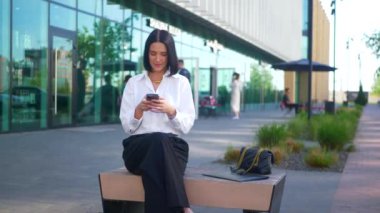 İş merkezinin yanındaki bankta oturan bir kadın telefonda konuşuyor. Ofis çalışanı, yönetmen, resmi kıyafetler. Beyaz gömlekli ve siyah pantolonlu bir kadın bankta cep telefonuyla sohbet ediyor ve caddenin karşısında gülümsüyor. Gülümseyen iş adamı oturuyor. 