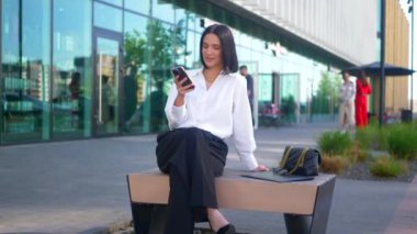 İş merkezinin yanındaki bankta oturan bir kadın telefonda konuşuyor. Ofis çalışanı, yönetmen, resmi kıyafetler. Beyaz gömlekli ve siyah pantolonlu bir kadın bankta cep telefonuyla sohbet ediyor ve caddenin karşısında gülümsüyor. Gülümseyen iş adamı oturuyor. 