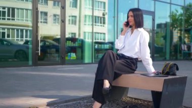 İş merkezinin yanındaki bankta oturan bir kadın telefonda konuşuyor. Ofis çalışanı, yönetmen, resmi kıyafetler. Beyaz gömlekli ve siyah pantolonlu bir kadın bankta cep telefonuyla sohbet ediyor ve caddenin karşısında gülümsüyor. Gülümseyen iş adamı oturuyor. 