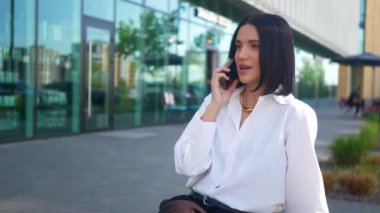 İş merkezinin yanındaki bankta oturan bir kadın telefonda konuşuyor. Ofis çalışanı, yönetmen, resmi kıyafetler. Beyaz gömlekli ve siyah pantolonlu bir kadın bankta cep telefonuyla sohbet ediyor ve caddenin karşısında gülümsüyor. Gülümseyen iş adamı oturuyor. 