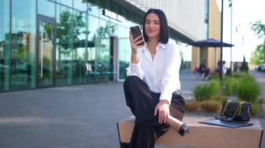 İş merkezinin yanındaki bankta oturan bir kadın telefonda konuşuyor. Ofis çalışanı, yönetmen, resmi kıyafetler. Beyaz gömlekli ve siyah pantolonlu bir kadın bankta cep telefonuyla sohbet ediyor ve caddenin karşısında gülümsüyor. Gülümseyen iş adamı oturuyor. 