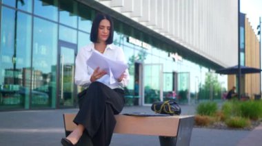 Kadın iş merkezinin yakınındaki belgelere bakıyor. Ofis çalışanı, yönetmen, resmi kıyafetler. Beyaz tişörtlü ve siyah pantolonlu kadın menkul kıymetlere bakıyor ve caddenin ortasında gülümsüyor. İş gülümseyen girişimci.