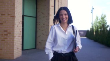 İş kadını yürüyor, gülümsüyor ve iş merkezinin yakınındaki kameraya bakıyor. Ağır çekim, ofis çalışanı, yönetmen, resmi kıyafet. Beyaz gömlekli ve siyah pantolonlu bir kadın caddenin ortasında gülümsüyor. İş gülümseyen girişimci.