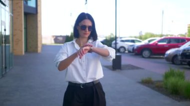 İş kadını yürüyor, gülümsüyor ve iş merkezinin yakınındaki kameraya bakıyor. Ağır çekim, ofis çalışanı, yönetmen, resmi kıyafet. Beyaz gömlekli ve siyah pantolonlu bir kadın caddenin ortasında gülümsüyor. İş gülümseyen girişimci.