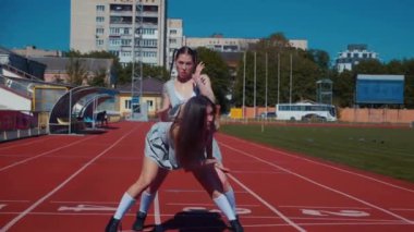 Stadyum pistinde neşeyle dans eden iki ateşli kadın. Yavaş çekim. Kısa etekli ponpon kızlar koşu pistinde mutlu bir şekilde dans ediyorlar. İki kız spor pistinde baştan çıkarıcı bir şekilde dans ediyor. Kadınlar enerjik bir şekilde dans ediyor 