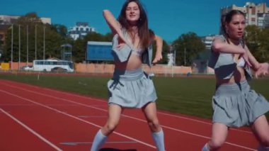 Stadyum pistinde neşeyle dans eden iki ateşli kadın. Yavaş çekim. Kısa etekli ponpon kızlar koşu pistinde mutlu bir şekilde dans ediyorlar. İki kız spor pistinde baştan çıkarıcı bir şekilde dans ediyor. Kadınlar enerjik bir şekilde dans ediyor 