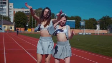 Stadyum pistinde neşeyle dans eden iki ateşli kadın. Yavaş çekim. Kısa etekli ponpon kızlar koşu pistinde mutlu bir şekilde dans ediyorlar. İki kız spor pistinde baştan çıkarıcı bir şekilde dans ediyor. Kadınlar enerjik bir şekilde dans ediyor 