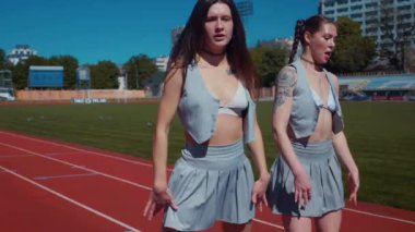 Stadyum pistinde neşeyle dans eden iki ateşli kadın. Yavaş çekim. Kısa etekli ponpon kızlar koşu pistinde mutlu bir şekilde dans ediyorlar. İki kız spor pistinde baştan çıkarıcı bir şekilde dans ediyor. Kadınlar enerjik bir şekilde dans ediyor 