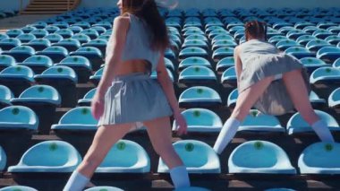 Stadyum pistinde neşeyle dans eden iki ateşli kadın. Yavaş çekim. Kısa etekli ponpon kızlar koşu pistinde mutlu bir şekilde dans ediyorlar. İki kız spor pistinde baştan çıkarıcı bir şekilde dans ediyor. Kadınlar enerjik bir şekilde dans ediyor 