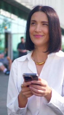 İş merkezinin yanındaki bankta oturan bir kadın telefonda konuşuyor. Dikey. Ofis çalışanı, yönetmen, resmi kıyafetler. Beyaz gömlekli ve siyah pantolonlu bir kadın bankta cep telefonuyla sohbet ediyor ve caddenin karşısında gülümsüyor. Gülümseyen iş