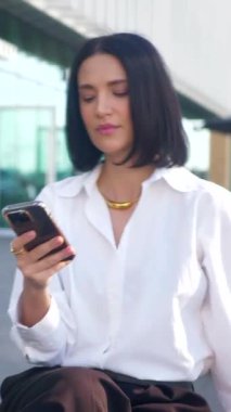 İş merkezinin yanındaki bankta oturan bir kadın telefonda konuşuyor. Dikey. Ofis çalışanı, yönetmen, resmi kıyafetler. Beyaz gömlekli ve siyah pantolonlu bir kadın bankta cep telefonuyla sohbet ediyor ve caddenin karşısında gülümsüyor. Gülümseyen iş