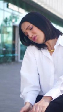 İş merkezinin yanındaki bankta oturan bir kadın telefonda konuşmaktan memnun değil. Dikey. Şehirdeki bir bankın ortasında iş telefonu konuşması yapan başarılı bir kadın. Oturan kadın telefonda konuşuyor. Beyaz gömlekli kadın.