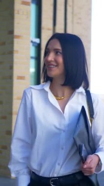 İş kadını yürüyor, gülümsüyor ve iş merkezinin yakınındaki kameraya bakıyor. Dikey, ofis çalışanı. Beyaz gömlekli ve siyah pantolonlu bir kadın caddenin ortasında gülümsüyor. İş gülümseyen girişimci.