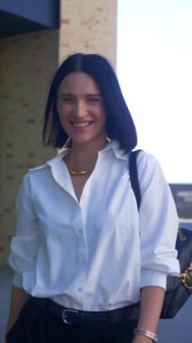 İş kadını yürüyor, gülümsüyor ve iş merkezinin yakınındaki kameraya bakıyor. Dikey, ofis çalışanı. Beyaz gömlekli ve siyah pantolonlu bir kadın caddenin ortasında gülümsüyor. İş gülümseyen girişimci.