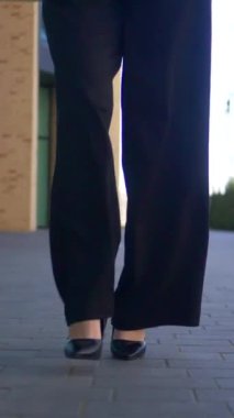 İş kadını yürüyor, gülümsüyor ve iş merkezinin yakınındaki kameraya bakıyor. Dikey, ofis çalışanı. Beyaz gömlekli ve siyah pantolonlu bir kadın caddenin ortasında gülümsüyor. İş gülümseyen girişimci.