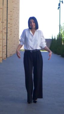 Dikey video. İş kadını iş merkezinin yakınında dans edip gülümsüyor. Ofis çalışanı, yönetmen, resmi kıyafetler. Beyaz gömlekli ve siyah pantolonlu bir kadın işi bittikten sonra caddenin ortasında dans ediyor. İş gülümseyen girişimci.