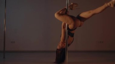 Woman in leather underwear gracefully pole dancing in a dimly lit room. Elegant female in a bikini performer doing a seductive dance on a pole. Lady showing her strength and flexibility on pole. Sensual dance performance by a woman on a metal pole