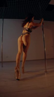Deri iç çamaşırlı kadın loş bir odada zarif bir şekilde dans ediyor. Dikey video. Bikini giyen bir kadın direğin üzerinde baştan çıkarıcı bir dans yapıyor. Bayan direkte gücünü ve esnekliğini gösteriyor..