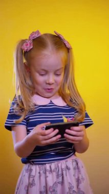 Güneş gözlüğü takan küçük bir kız cep telefonuyla oyun oynuyor. Dikey video, güneş gözlüklü genç bir kız cep telefonuna bakıyor ve mobil bir sanal oyun oynarken mutlu bir şekilde gülüyor. Çocuk seviniyor, elinde bir cep telefonu. 