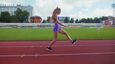 Genç bir kadın antrenmana hazırlanıyor. Spor meraklısı koşu parkurunda topallıyor. Hareketli kadın egzersiz öncesi esneme hareketleri yapıyor. Koşucu sporcu esneme hareketleriyle ısınıyor. Dişi koşucu ön koşular yapıyor