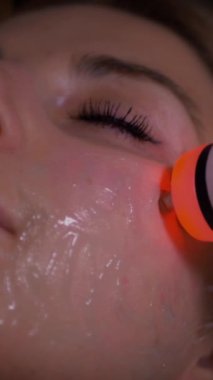 Dikey olarak, genç bir kadın spa salonunda profesyonel donanım titreşim yüz masajı yaptırıyor. Güzel kız yüzüne rahatlatıcı bir masaj yaptırıyor. Yakın plan çekim. Lüks profesyonel masaj anlayışı. Ağır çekimde çekmek.