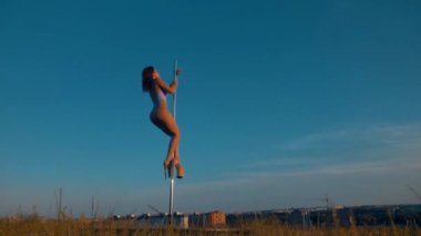 Kadın zarif bir şekilde gökyüzü zemininde dans ediyor. Polonyalı dansçı dışarıda güç ve esneklik gösteriyor. Açık gökyüzünün altında zarif bir direk dansı. Kadın dansçı, mavi bir arka plana karşı direkte hassas hareketler yapıyor.. 