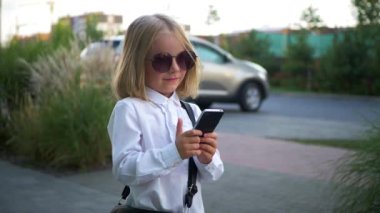 Güneş gözlüklü küçük mutlu bir kız cep telefonunu tutarken gülümsüyor. Genç bir kız şık güneş gözlüğü takıyor, akıllı telefonuyla oynuyor ve çok mutlu. Güneş gözlüklü neşeli bir kız, telefonuyla anları yakalıyor..
