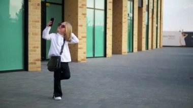 Genç bir kız elinde telefonuyla mutlu, komik bir şekilde dans ediyor ve kulaklıklarıyla müziğin tadını çıkarıyor. Genç bir iş kadını, iş merkezinin yakınında telefonu elinde tutarak mutlu olur. Kulaklık takan şık bir çocuk., 