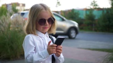 Güneş gözlüklü küçük mutlu bir kız cep telefonunu tutarken gülümsüyor. Genç bir kız şık güneş gözlüğü takıyor, akıllı telefonuyla oynuyor ve çok mutlu. Güneş gözlüklü neşeli bir kız, telefonuyla anları yakalıyor..