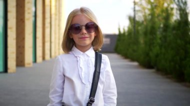 Beyaz gömlekli ve siyah pantolonlu genç bir kız kaldırımda duruyor, gülümsüyor ve kameraya bakıyor. Şehir kaldırımında kendine güvenen bir duruşu olan beyaz bluz ve siyah pantolon giyen küçük bir kız. Beyaz gömlek giyen neşeli bir kız 