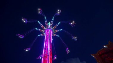 Mavi gökyüzüne karşı parlayan ışıklarla süslenmiş canlı bir eğlence gezisi. Luna Parkı. Lunaparktaki dönme dolap. Ekstrem atlıkarınca sevinçle dönüyor. Güneşli bir gökyüzünde yeşil ışıklarla parıldayan neşeli bir eğlence..